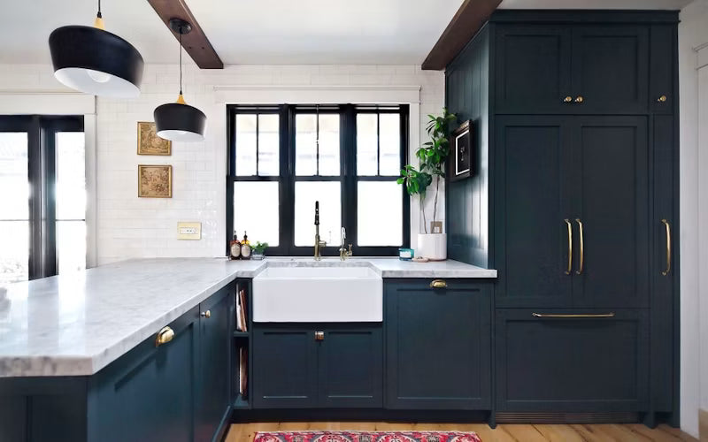 A modern kitchen featuring dark navy blue cabinets with brass handles, a white farmhouse sink, a white marble countertop, and black pendant lights.