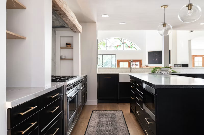 A modern kitchen interior with black cabinetry, stainless steel appliances, white countertops, wood flooring, and pendant lights. The scene shows the kitchen layout rather than a single isolated product, and no person or animal is present.