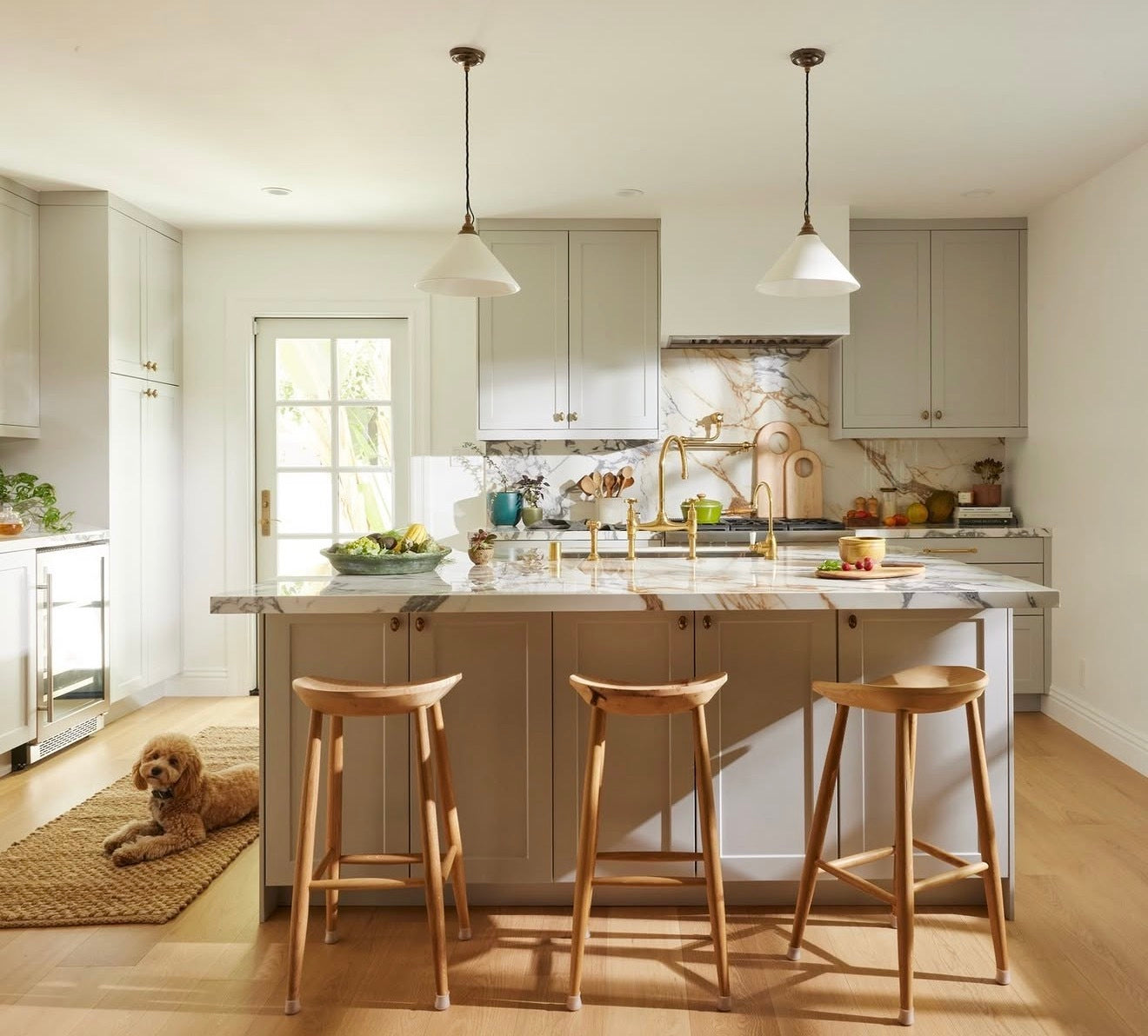 A bright, modern kitchen with light gray cabinets, a marble island, three wooden bar stools, and a small dog lying on a woven rug on the floor.