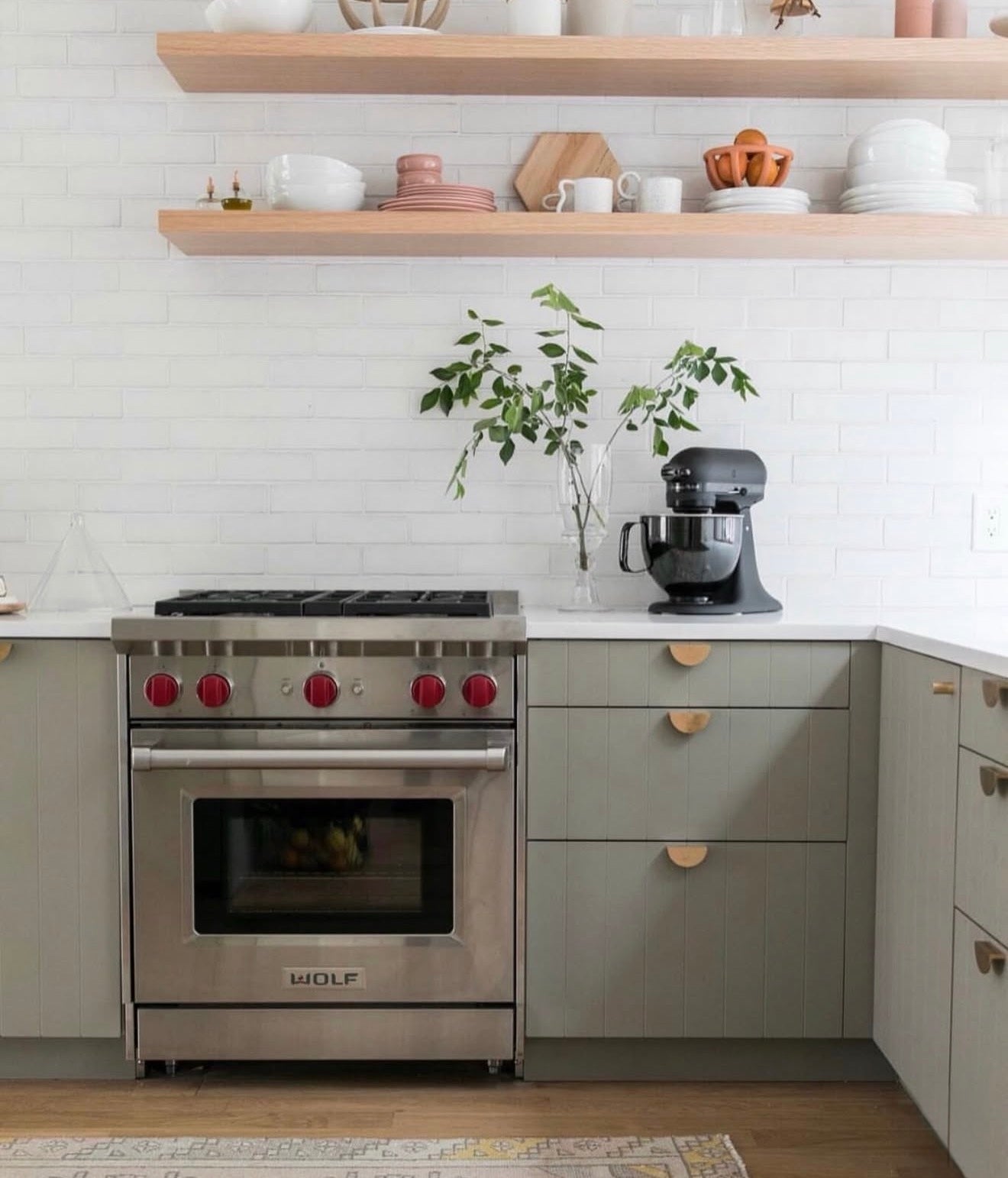 A modern kitchen scene featuring a stainless steel Wolf gas range with red control knobs, set in sage green cabinets with light wood drawer pulls. Above the stove, light wood floating shelves hold white and pink ceramic dishes, with a white subway tile backsplash behind.
